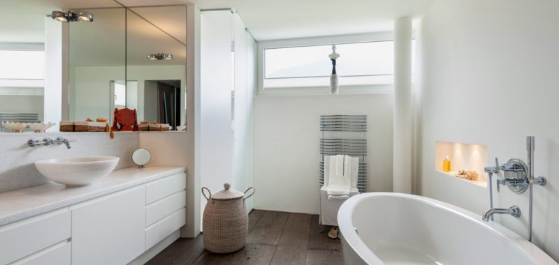 Spacious and minimalist white bathroom with a freestanding bathtub, offering a serene ocean view through large windows.