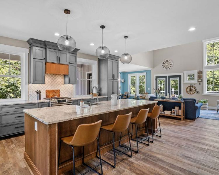 Contemporary kitchen featuring dark gray cabinets, wooden accents, and a spacious white island, surrounded by natural scenery visible through large windows.